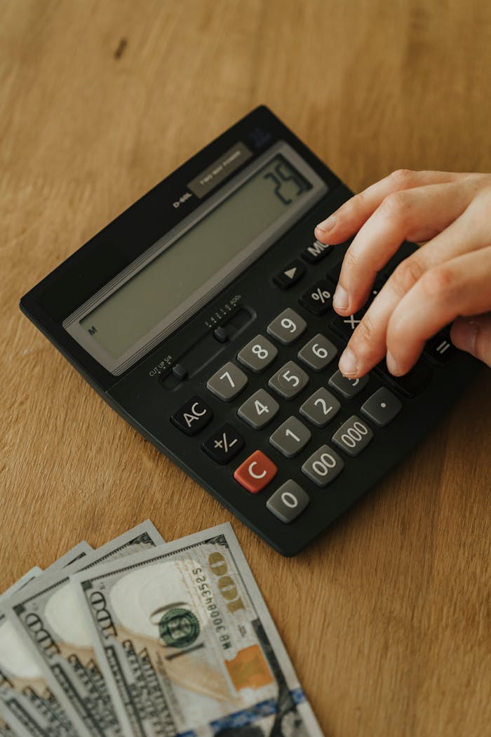 Hand using a calculator and counting hundred dollar bills on a wooden table, concept of personal finance management.